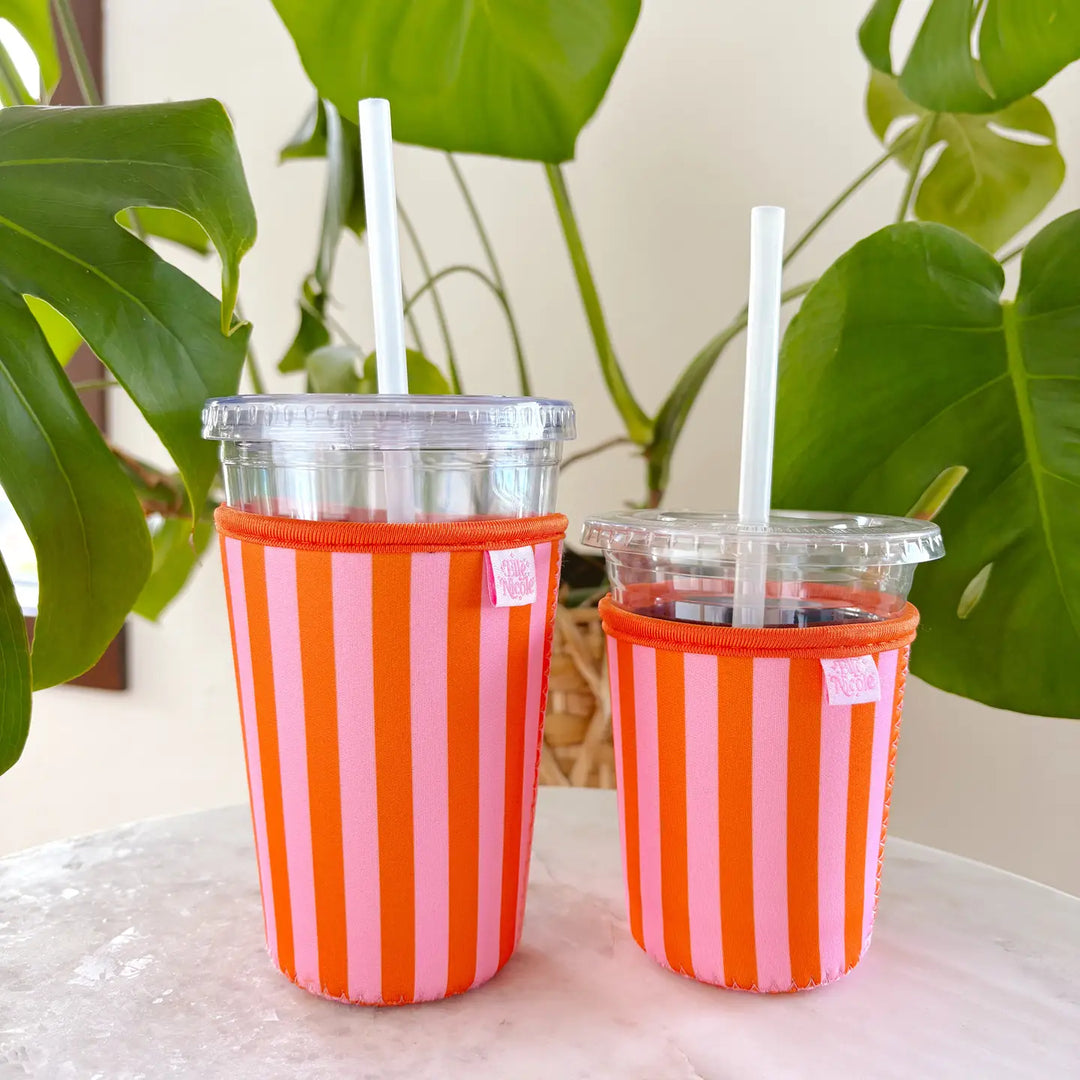 Two sizes of iced coffees in red and pink striped neoprene sip sleeves with green plant in background