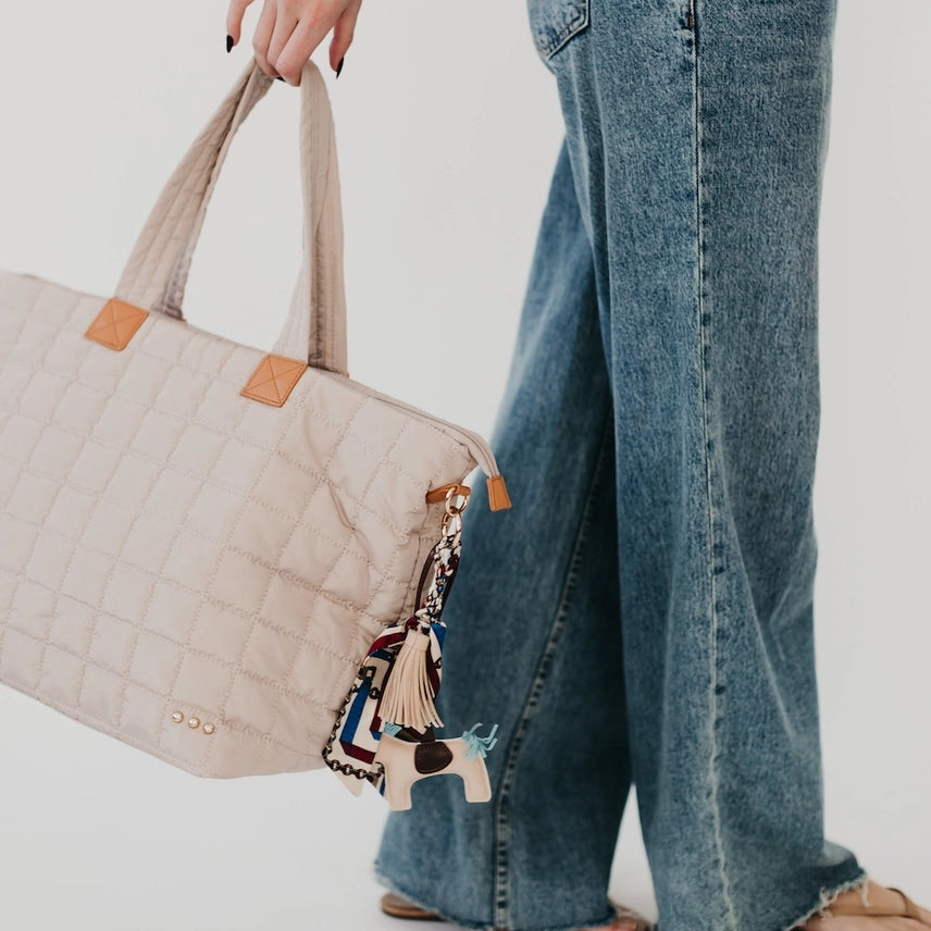 Secretariat Bag Charm with a horse pendant, mini silk scarf, and tassel, designed as a stylish add-on for handbags and totes. Available in tan or gray. Photo of the bag charm styled on the Day Dreamer Quilted Tote Bag. 