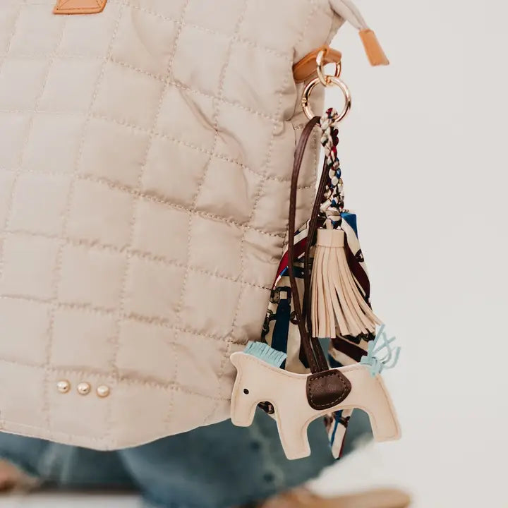 Secretariat Bag Charm with a horse pendant, mini silk scarf, and tassel, designed as a stylish add-on for handbags and totes. Available in tan or gray. Photo of the bag charm styled on the Day Dreamer Quilted Tote Bag.