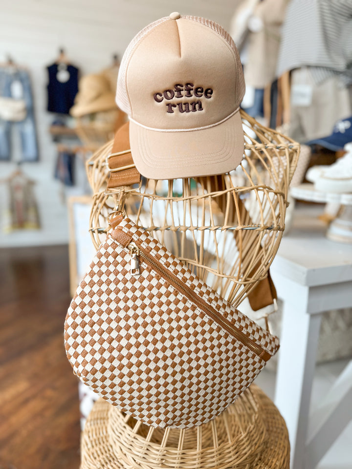 Checkered brown and white woven Maddox crossbody belt bag with adjustable strap pictured on a wicker mannequin with a hat that says 'coffee run'.
