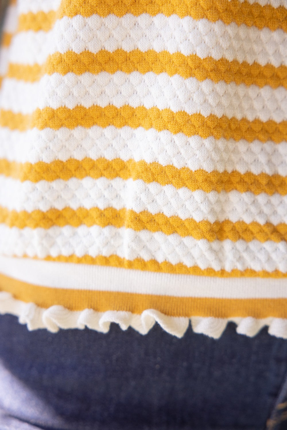 Close-up of hem ruffle trim detail of mustard and white striped knit short sleeve blouse with ruffle trim, styled as a casual work top for women.