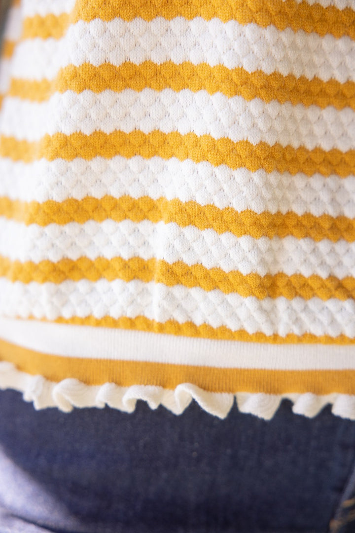 Close-up of hem ruffle trim detail of mustard and white striped knit short sleeve blouse with ruffle trim, styled as a casual work top for women.